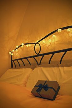 Gift surprise concept. Valentine’s day date concept. Cutely wrapped gift box with blue bow placed on a comfortable bed decorated with garland of Christmas lights around the wrought iron headboard. Romantic atmosphere concept.