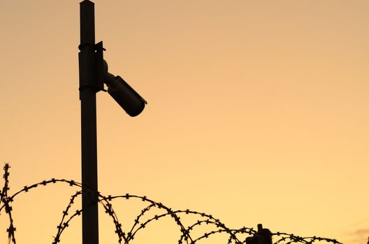Silhouette of the Security camera on the pole on sunset.Observation of the perimeter of the protected area with barbed wire.Space for text