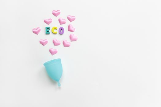 Turquoise menstrual cup on white background with hearts and word Eco from colorful wooden letters. Concept zero waste, savings, minimalism. Feminine hygiene product flat lay, copy space. Horizontal.