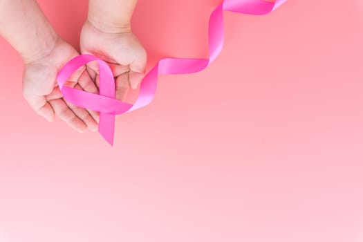 Woman hands holding pink ribbon breast cancer on pink background with copy space for text. Flat lay, top view.