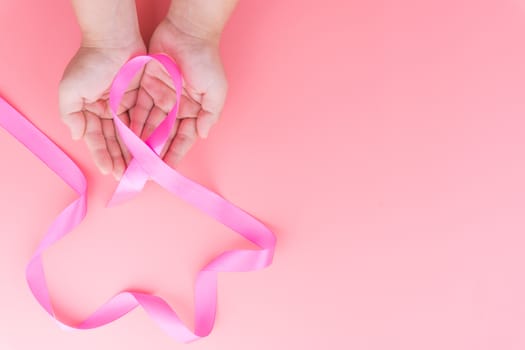 Woman hands holding pink ribbon breast cancer on pink background with copy space for text. Flat lay, top view.