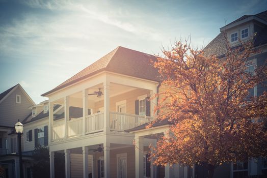 Bright fall foliage near typical two story country-style residential house in suburbs Dallas. White painted porch patio with banister, dormer roof and outdoor ceiling fan on second floor