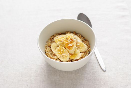 White ceramic bowl of oatmeal with banana slices and honey, served with the spoon on white table background. Viewed from high angle in closeup