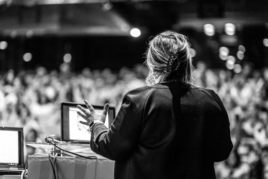 Female speaker giving a talk on corporate business conference. Unrecognizable people in audience at conference hall. Business and Entrepreneurship event. Black and white image.