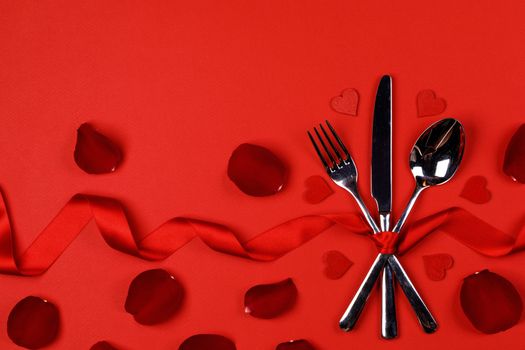 Cutlery set tied with silk ribbon rose flower petals and hearts on red background Valentine day dinner concept