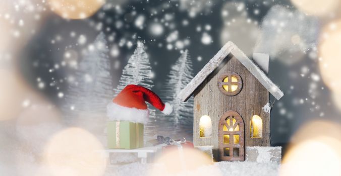 Christmas card with small glowing decorative house , gifts and Santa Claus cap on sleigh on snow over fairy tale winter fir forest background and falling snow . New Year design postcard