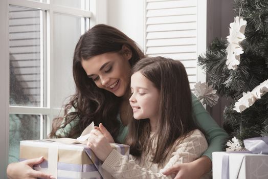 Happy mother and her little daughter unpack gift box near the Christmas tree at home