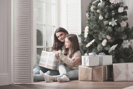 Happy mother and her little daughter unpack gift box near the Christmas tree at home