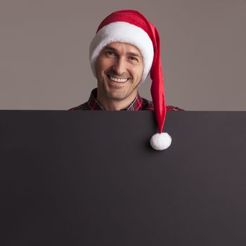 Man wearing santa christmas hat holding and pointing blank billboard or placard sign copy space for text advertising