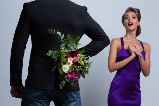 Man brings flowers to woman and hiding them behind back, womanin evening dress is surprised and happy