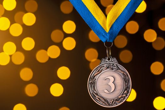 Third placed runner up bronze medallion on a ribbon hanging against a blue background with golden bokeh for a celebration of the win in a sporting competition or championship