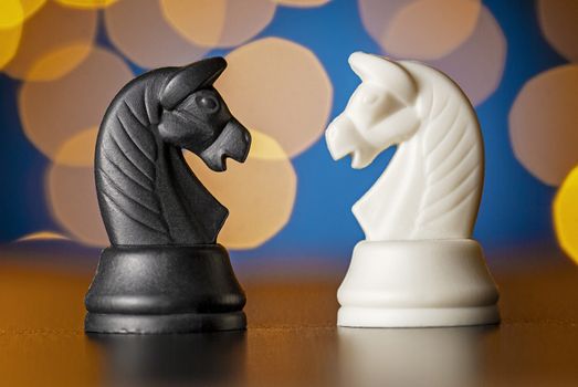 Two horse chess pieces in black and white standing on a wooden table facing each other against a colorful golden background bokeh effect