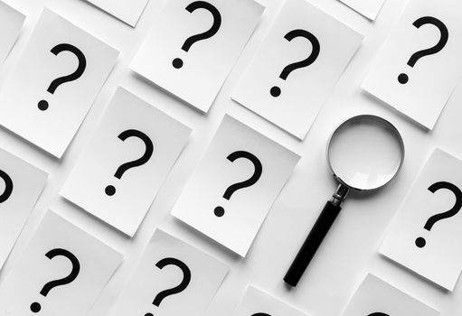 Diagonally orientated question marks printed on white paper cards and a magnifying glass in a neat background pattern viewed from overhead