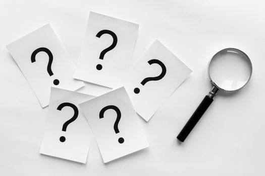 Question marks printed on white cards lying on a white background with a magnifying glass in a conceptual image