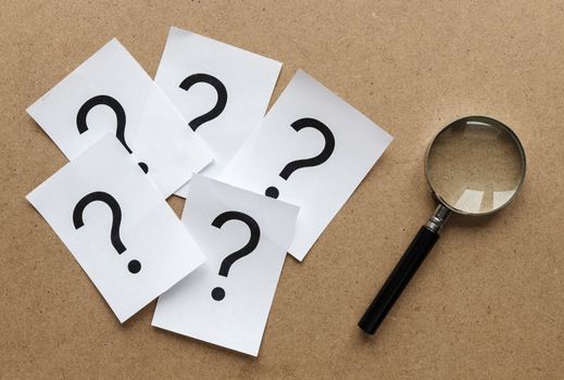 Question marks printed on white cards lying on a wooden background with a magnifying glass in a conceptual image