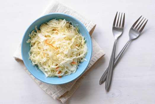 Blue bowl of shredded chopped pickled cabbage and carrot. Served with two forks on folded grey fabric towel and viewed from above on white table surface background