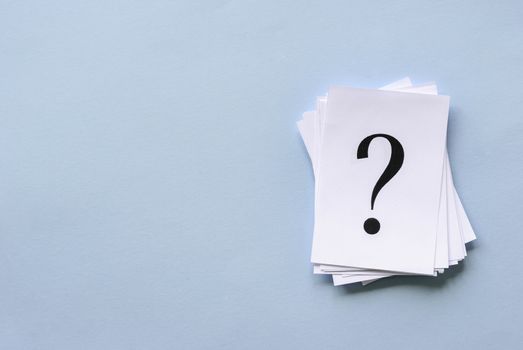 Pile of stacked question marks printed on sheets of white paper or signs arranged to the side on a blue background with copy space in a conceptual image