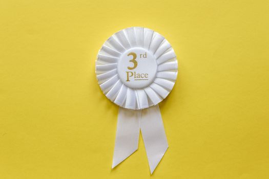 3rd place white ribbon rosette with gold text for the runner up in a competition or race on a yellow background with copy space