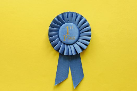 Blue ribbon rosette for the First Placed Winner of a race, championship or competition over a yellow background with copy space