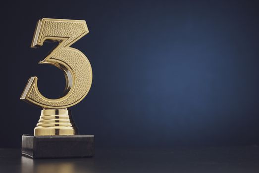 Sharp close up on single isolated gold colored plastic statue of number 3 over blue and black background