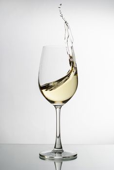 White wine splashing up the side of an elegant long stemmed wineglass in a freeze motion image on a reflective surface against a white background