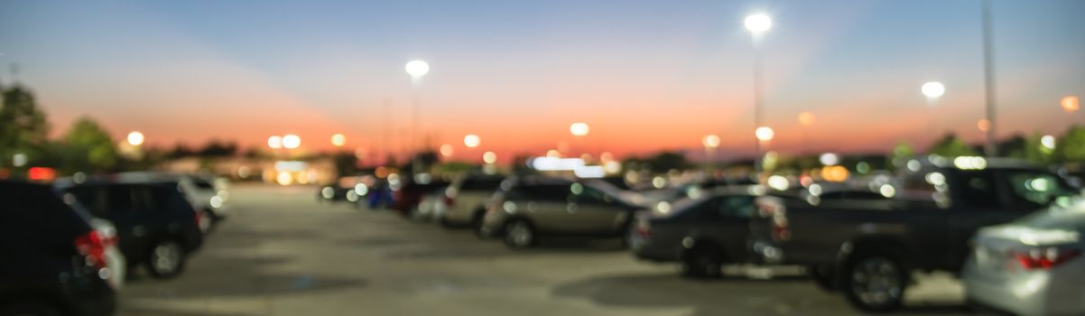 Panorama view abstract blurred parking lot of modern shopping center in Houston, Texas, USA. Exterior mall complex with row of cars in outdoor uncovered parking, bokeh light poles in background