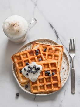 Delicious homemade baked belgian waffles with greek yogurt, blueberries and cappuccino on white marble background. Perfect breakfast with copy space for text or design. Top view or flat lay. Vertical