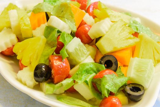 Close-up fresh dish of mixed organic fruits salad with pineapple, cucumber, apple, cherries tomatoes, lettuce, carrot, olive. Colorful and healthy fiber in white leaf shape plate.