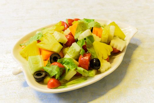 Fresh dish of mixed organic fruits salad with pineapple, cucumber, apple, cherries tomatoes, lettuce, carrot, olive. Colorful and healthy fiber in white leaf shape plate.