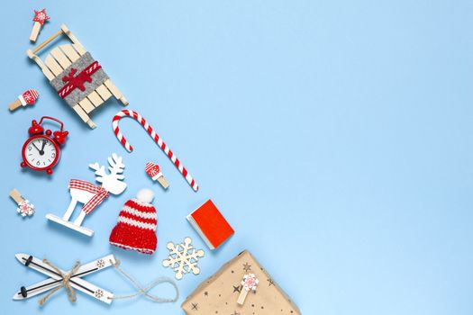 Corner creative Christmas composition. Candy cane, gift, sled, deer, hat, alarm clock, ski, clothespins, book, wooden snowflakes on blue background, copy space. Minimal style. Top view.