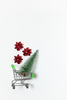 Christmas fir and red bows for gift in toy shopping cart on white background, copy space. New Year, sales, online shopping concept. Vertical, flat lay. Minimal style. Top view. For social media.