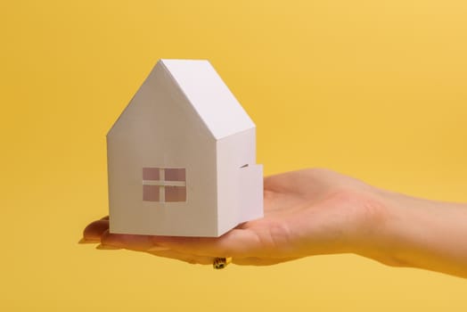 White family paper house in man hand on yellow background paper. Minimalistic style. Copy space. View from above. Vertical orientation