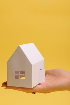 White family paper house in man hand on yellow background paper. Minimalistic style. Copy space. View from above. Vertical orientation
