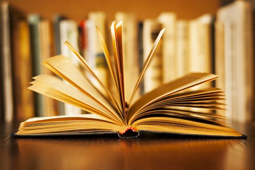 Hardcover book lying open on its cover with the pages fanned above it and a row of books on a bookshelf visible behind, shallow dof