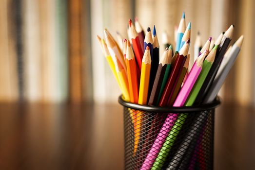 Wire desk tidy full of coloured pencils standing on a wooden table in front of a bookshelf full of books with shallow dof and copyspace