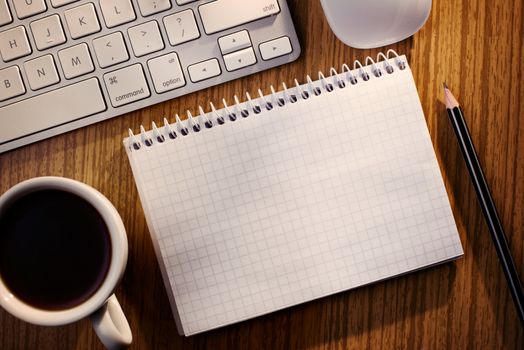 Open notebook with blank white lined pages with a cup of black coffee alongside a computer keyboard on a wooden desk, overhead view
