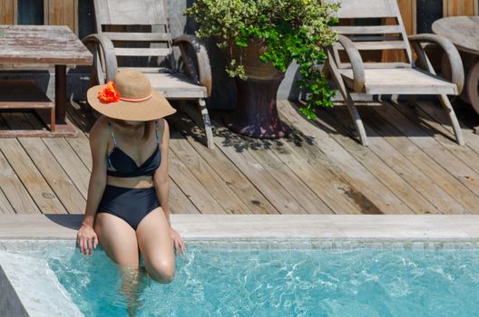 Beautiful young woman sitting by the pool wearing a brown hat.
