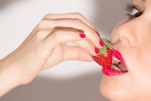 Close up of a woman with red lips and nails, biting a ripe strawberry