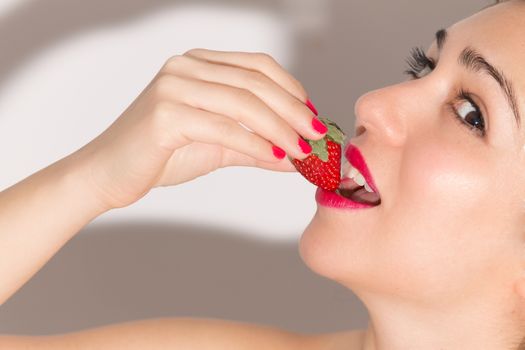 Close up of a woman with red lips and nails, biting a ripe strawberry