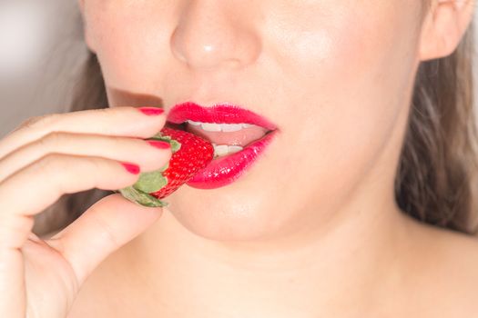 Close up of a woman with red lips and nails, biting a ripe strawberry