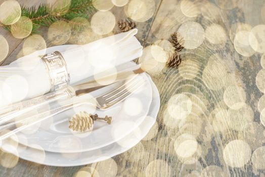 Christmas table: silver knife, fork and linen napkin lie on the white dinner plate, as well as green spruce branch and cones which is located on a old wooden table, with space for text