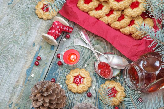 Beautiful Christmas card: Sweet Christmas cookies with jam, fir branches and cones, cranberries, red burning candle and the bobbin with decorative ribbon on the old wooden table, with copy-space