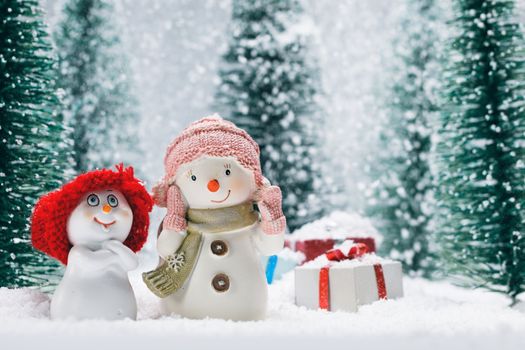Two small cute snowmen friends with gifts in forest under falling snow