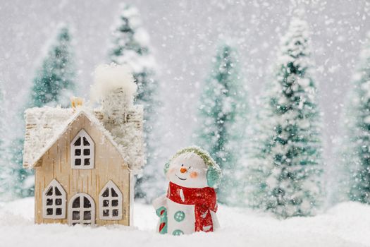 Small decorative snowman near wooden house in fir forest under falling snow