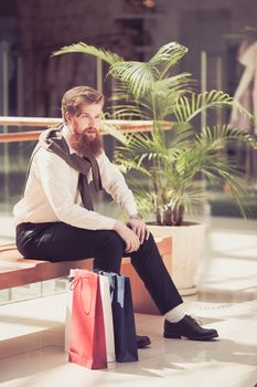 Smiling redhead bearded male customer in trendy wear sitting in store with bags after shopping and buying purchases