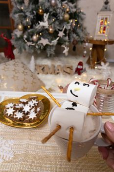 Marshmallow snowman floats in a delicious mug of hot chocolate at Christmas time with a plate of iced gingerbread stars.  Festive decorated tree and Christmas ornmanets in background