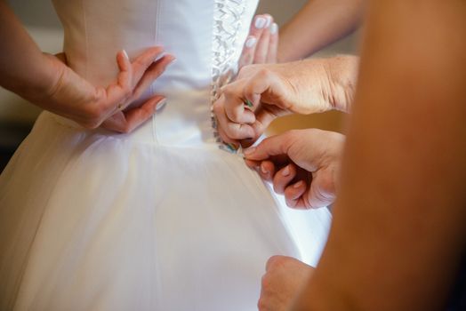 Bridesmaid helping slender bride lacing her wedding white dress, buttoning on delicate lace pattern with fluffy skirt on waist. Morning bridal preparation details newlyweds. Wedding day moments, wear.