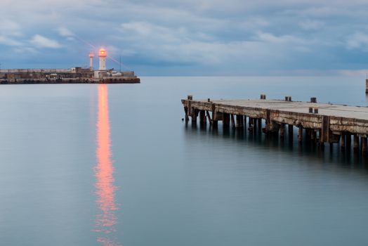 The bright light of the lighthouse on the seashore, right in the frame of the pier at sunset