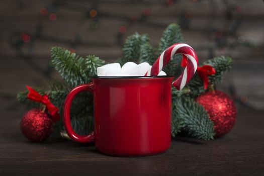 Cocoa in red mug with marshmallows candy cane fir tree branches and red baubles on dark wooden background with bokeh lights