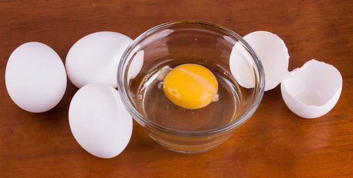 Broken egg in a glass bowl with whole eggs and shell on wood table